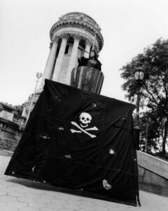 A towering structure showcases ornate columns in a classical style, set in a public space with a cobblestone path. In the foreground, a dramatic setup features a tattered black sheet with a skull and crossbones motif. A figure stands prominently in a wooden structure above it, suggesting a dynamic, thematic performance. Lush trees complete the scene.