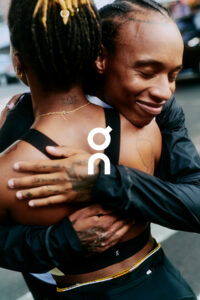 Two individuals are embracing warmly, creating a sense of connection and affection. Both have their eyes closed in contentment. The person facing away has a lotus tattoo on the back of their neck, and a gold chain is visible. They wear athletic apparel, suggesting an active lifestyle. Their hairstyles and tattoos add individual expressions to the scene.