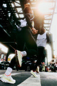 Two individuals in athletic gear are running under an elevated train track. Sunlight filters through the structure, casting dramatic shadows on the ground. They wear bright sneakers, and the urban setting includes cars and pedestrians in the background. The scene captures a sense of motion and energy amidst the bustling city environment.