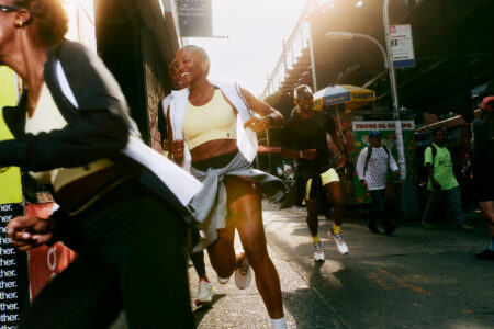 A group of people energetically run along a vibrant city street in the bright sunlight. The atmosphere is lively, with various individuals in motion, dressed in athletic attire. The urban setting features buildings, street signs, and shadows cast by the strong sun. Passersby observe the scene, contributing to the dynamic and bustling ambiance of the area.