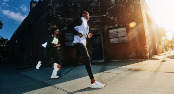 Two individuals are jogging down a sunlit urban street, with the sun casting a warm glow across the scene. They are dressed in athletic gear, including sleek black leggings and jackets, and their white sneakers contrast with the pavement. The backdrop features a rugged brick building, adding an industrial feel to the lively, energetic morning activity.