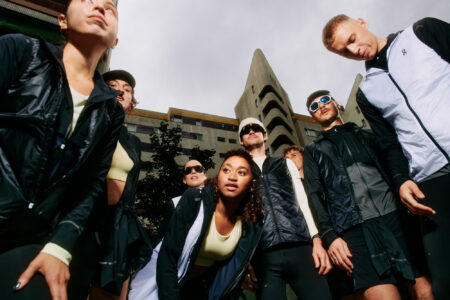 A group of fit, young adults stands collectively outdoors, sporting dark athletic wear. They look upward, illuminated by natural light, as modern buildings rise behind them. The sky appears overcast, casting a soft light. Among the group are men and women, some wear sunglasses, all exuding a sense of readiness and confidence. Their expressions are serious yet relaxed.