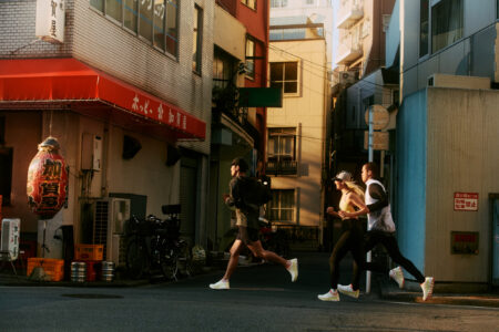 Three individuals jog along a narrow urban street, surrounded by buildings with a mix of modern and traditional architecture. A large decorative object hangs near the entrance of an establishment, adding cultural character. The street is quiet and bathed in warm daylight, casting soft shadows. Bicycles are parked nearby, contributing to the everyday city scene.