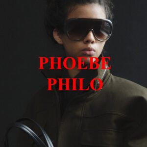 A person stands against a dark background, exuding a confident and stylish demeanor. They wear oversized sunglasses that dominate their facial features. Dressed in a high-collared, dark green jacket, the individual appears modern and edgy. Their hair is styled back, adding to the overall sleek look, while they hold a handle, possibly of a bag or accessory, in their hand.