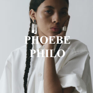 A person with long, dark hair styled in braids gazes thoughtfully. They are wearing a crisp white shirt, creating a minimalist and elegant appearance. Large geometric silver earrings and a matching ring add a touch of modern sophistication. Their hand rests on their face, contributing to a contemplative mood, set against a plain, light-colored background.