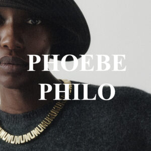 A person with a serious expression is seen wearing a wide-brimmed hat and a textured, dark sweater. The upper garment features a chunky gold necklace with unique, sculptural links. The background is plain and minimalistic, highlighting the individual's strong, contemplative gaze. The overall aesthetic is sophisticated and understated, emphasizing subtle elegance and style.