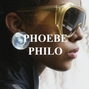 A close-up profile showcases a person wearing oversized, translucent yellow sunglasses with a reflective surface. The individual sports a dark, sleek garment and a distinctive large, glossy metallic circular earring. The lighting highlights the texture and color of their skin. The background is blurred, adding depth and focusing attention on the face and accessories.