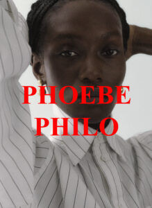 A person gazes intently, wearing a white shirt with thin, dark vertical stripes. The shirt has a high collar and is buttoned up. The person has dark hair styled in a neat manner and wears a small, gold earring. The background is plain and light, emphasizing the subject's thoughtful expression and poised posture.