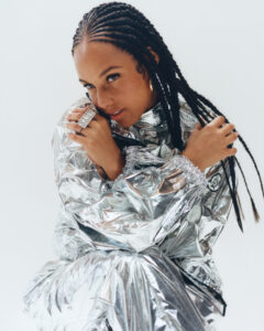 A person with long braided hair is wearing a shiny metallic outfit. They hold their hands close to their face, showcasing a large, ornate ring. The background is a simple white, highlighting the reflective quality of the clothing. The person's expression is soft and confident, with warm lighting enhancing the overall vibrant, futuristic aesthetic.