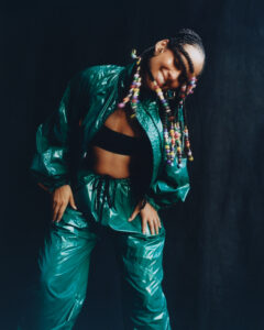 A person is joyfully leaning forward, wearing a shiny green tracksuit with a black top underneath. Their hair is styled in braids adorned with colorful beads, adding a playful touch. The background is a dark, plain backdrop that contrasts with the vibrancy of their outfit. Their wide smile and dynamic pose convey energy and freshness.