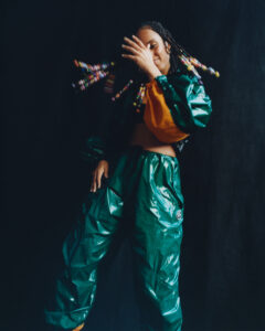 A person stands against a dark backdrop, wearing a vibrant, shiny green and orange outfit. Their hair is adorned with colorful beads, adding an eclectic and playful element to their look. The person's pose is dynamic, with one arm raised, partially concealing their face, exuding a sense of energy and movement. The overall composition is bold and expressive.