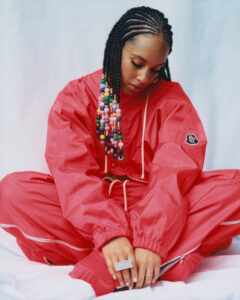 A person sits cross-legged, wearing a vibrant red tracksuit with cinched cuffs and visible stitching. Their hair is styled in intricate braids adorned with colorful beads, adding a playful touch. They look downward in a contemplative manner. A ring adorns their finger, and the overall composition evokes a sense of relaxed confidence against a light background.