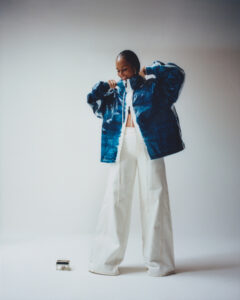 A person stands confidently in a minimalist setting, wearing a reflective blue jacket with a high collar and wide-legged white pants. The individual, whose hair is styled neatly, appears to be adjusting the jacket while smiling. The lighting creates a soft contrast, enhancing the modern, stylish atmosphere. A small, metallic object is positioned at their feet.