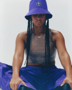 A person with long braided hair sits in a relaxed pose. They wear a sparkling, sleeveless top and vibrant purple pants. A matching purple bucket hat is stylishly placed on their head. The overall atmosphere is casual yet fashionable, with a focus on modern streetwear. The background is minimalistic, emphasizing the person's attire and presence.