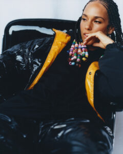 A person with braided hair adorned with colorful beads reclines on a glossy black chair. They wear a shiny, black jacket with a vibrant yellow lining. Their expression appears contemplative, with one hand resting near the chin and eyes gazing thoughtfully into the distance. The attire and relaxed pose convey a sense of modern, casual elegance.