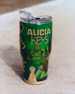 A vibrantly decorated tumbler stands on a rough surface. It features a green background adorned with sparkling embellishments creating intricate patterns. A skyline and bridge are artistically depicted, adding a sense of urban charm. The tumbler has a clear lid with a straw for drinking. Its overall aesthetic blends functionality with decorative flair, making it visually striking.
