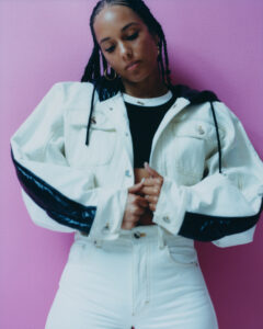 A person with braided hair is wearing a white jacket with black details and high-waisted pants. They stand against a vibrant pink background. Their head is tilted slightly downward, showing a calm expression. Large hoop earrings and a subtle necklace complete the look, blending casual style with a hint of elegance and attitude.