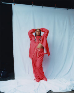A person dressed in a vibrant red tracksuit stands against a minimalist white backdrop. They express confidence with relaxed posture, arms raised above the head. The outfit is detailed with colorful beads in the hair, adding a playful touch. The setting is a studio with fabric draped on the floor, creating a casual yet striking scene.