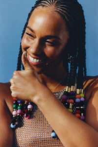 A person with intricately braided hair featuring colorful beads smiles warmly, giving a thumbs-up. The background is a soft sky blue, which highlights the subject's radiant complexion. They wear a sparkling, studded top that catches the light. The overall mood is joyful and confident, with a sense of playful positivity emanating from the expression and vibrant details.