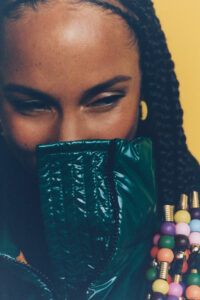 A person is partially concealed behind a glossy, dark green garment, capturing a playful or enigmatic demeanor. They have tightly braided hair with colorful beads, adding a vibrant flair. The background is a solid yellow, enhancing the overall warmth. The visible eyes are beautifully accentuated, focusing intently, while the scene conveys a sense of mystery and style.