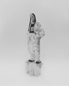In grayscale, a person stands confidently atop a textured pedestal, wearing a shiny, oversized metallic jacket and matching pants. The ensemble is complemented by a black top and dark shoes. Hair is styled in long braids, emphasizing an edgy, futuristic fashion statement amidst a minimalist backdrop that draws focus to both the attire and the stance.