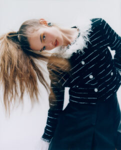 A person with long, flowing hair styled in a high ponytail tilts their head to the side. They wear a black, patterned outfit adorned with buttons and a white, fluffy collar. The makeup is bold, emphasizing their eyes, and they accessorize with large, circular earrings. The overall pose and fashion create a dynamic and stylish composition.