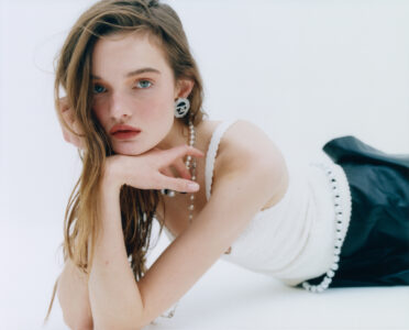 A person is lying down, gazing directly with an introspective expression. Long hair drapes naturally over one shoulder. They wear a textured top with thin straps and a dark skirt. Accessories include pearl jewelry and statement earrings. The background is soft and minimal, emphasizing an elegant and contemplative mood. The overall tone is serene and stylish.