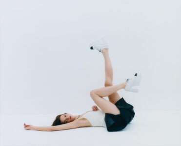 A woman lies on her back, her body forming a playful pose against a plain background. She wears a white top and a dark skirt, with one leg raised and bent at the knee. Her feet are clad in chunky white heels. Her expression is relaxed, with her head turned slightly to the side, exuding a sense of casual elegance.