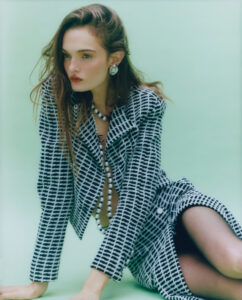 A person with long hair is poised against a soft green background. They wear a black and white checkered jacket with matching skirt, revealing a layered pearl necklace underneath. Large pearl earrings complement the attire. The person's gaze is thoughtful, directed off to the side. Their posture is relaxed yet confident, with one knee bent.