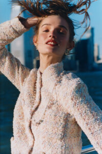 A person stands with a confident expression, hand in hair, against a backdrop of an urban skyline. They wear a textured, light-colored jacket with pearl accents and elegant earrings. The breeze tousles the hair, capturing a moment of poise and sophistication. The sun highlights the cityscape, adding warmth to the scene, evoking a sense of cosmopolitan elegance.
