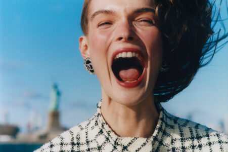 A woman with short hair is laughing joyfully against a clear blue sky. She is wearing a black and white checkered outfit and large decorative earrings. Her mouth is open wide, showing her teeth, and her eyes are closed in delight. In the blurred background, a well-known monument stands, adding a touch of iconic scenery.