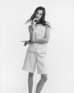 A woman poses with a relaxed yet thoughtful expression. She wears a textured, sleeveless top with large buttons and matching knee-length shorts. Her long hair flows gently, adding a sense of movement. Her left hand rests on her right arm, adorned with a bracelet. The monochrome style highlights the simplicity and elegance of her outfit and demeanor.