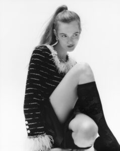 A person sits in a poised pose, with one knee bent. Their hair is styled in a high ponytail, and they are wearing an elegant, textured jacket with feather detailing. The outfit is completed with sheer, patterned knee-high socks and a leather skirt. The monochromatic scheme adds a classic, sophisticated touch, highlighting their confident expression.