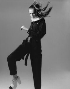 A person strikes a dynamic pose, dressed in a dark outfit with white accents. The fashion is bold, featuring a belt and a flowing hairstyle that adds motion. One leg is lifted, displaying white, chunky shoes. The monochrome palette enhances the dramatic and stylish aura, capturing a sense of movement and confidence against a plain backdrop.