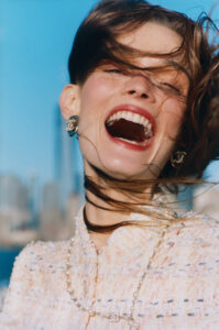 A woman with a big, joyful smile laughs openly in a dynamic, candid moment. Her hair is playfully tossed by the wind, and she wears elegant earrings and a tweed outfit adorned with a pearl necklace. The background reveals an urban skyline under a clear blue sky, highlighting a scene full of vibrant energy and happiness.