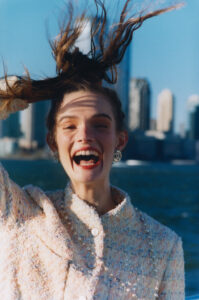A joyful woman with long hair held up high grins widely against a bright, urban backdrop. She wears a textured, pastel garment with buttons and jewel-like earrings. The skyline in the distance features tall buildings under a clear blue sky. Her animated expression and playful pose convey a sense of freedom and happiness on a sunny day.