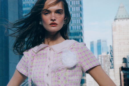 A woman stands in front of an urban backdrop with tall buildings. She wears a pastel-colored tweed outfit with a flower embellishment and pearl earrings. Her long hair flows in the breeze, and she gazes confidently forward. The sky is bright blue, contrasting with the modern architecture. The scene exudes a sense of style and sophistication.