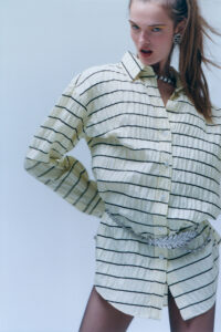 A person poses dynamically, exuding confidence, wearing a loose, striped button-down shirt accentuated by a sparkly belt. Their hair is styled in a high ponytail, revealing bold earrings and a thick, metallic necklace. The expression is fierce and engaging, with eyes accentuated and lips slightly parted, creating an edgy, fashion-forward aesthetic.