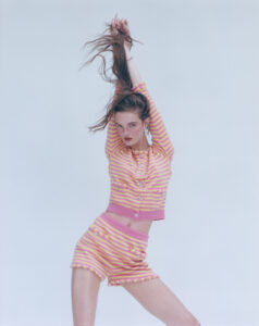 A person is posing dynamically against a plain backdrop, stretching their arms upwards while holding their long hair. They wear a coordinated outfit with pink and yellow horizontal stripes, featuring a fitted cardigan and matching shorts adorned with buttons and subtle fringe detailing. The overall composition is vibrant, conveying a sense of movement and playful elegance.