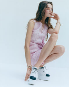 A young woman squats gracefully, wearing a sleeveless pink textured dress. Her long hair flows loosely over her shoulders. She accessorizes with a chunky gold bracelet. On her feet are striking white platform shoes with black toe caps and laces. She rests her chin on her hand, gazing calmly forward, in a minimalist setting.
