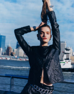 A person stands by a waterfront, lifting their hair with both hands. They're dressed in a black, quilted jacket and matching pants with a large metallic chain belt. The background reveals a cityscape with modern architecture and a boat in the water. The sky is blue with scattered clouds, creating a vibrant, urban atmosphere.