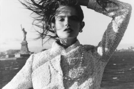 A woman stands with a hand in her hair, wearing a textured jacket and earrings, set against a breezy backdrop. Her hair blows in the wind, creating a dynamic and stylish scene. In the distance, the iconic statue adds a monumental presence, enhancing the timeless elegance of the composition with its classic black and white finish.