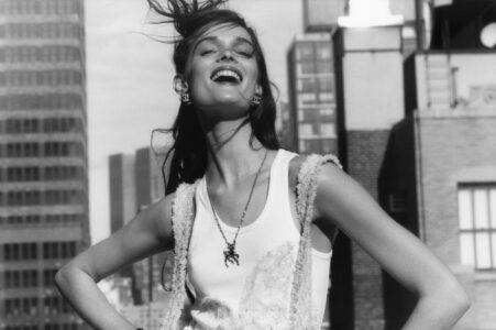 A joyful woman with wind-swept hair stands confidently in a cityscape. Her wide smile exudes happiness as she wears a stylish, textured tank top and a distinctive necklace. The monochrome background focuses on urban architecture, highlighting tall buildings. The scene suggests a vibrant, carefree moment, emphasizing fashion and outdoor energy amidst a bustling metropolis.