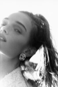 A softly focused black-and-white depiction captures an elegant woman’s profile. Her hair is styled in a ponytail, and she wears prominent, ornate earrings. The lighting creates gentle highlights and shadows, enhancing the serene, dreamy atmosphere. Her delicate expression and poised posture suggest an air of timeless sophistication and grace, reminiscent of classic portraiture from a bygone era.