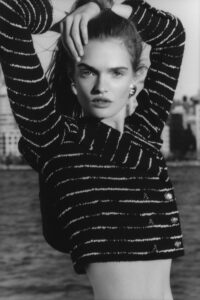 In a black-and-white scene, an individual stands gracefully before a waterfront, their hair pulled back. They wear a striped, buttoned top and striking earrings. Their arms are raised above their head, and the background showcases a blurred city skyline. The lighting emphasizes their features and creates a classic, timeless atmosphere, blending fashion with urban elements.