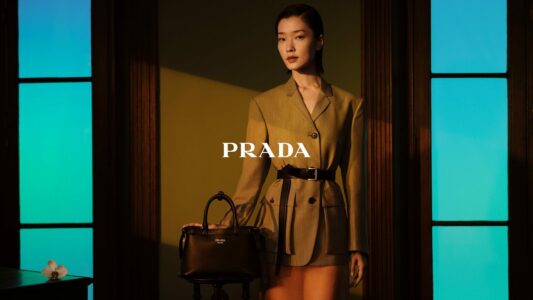 A poised individual stands confidently, dressed in a stylish tan blazer with a black belt, against a backdrop of rich, dramatic lighting. They hold a sleek black handbag, embodying elegance and sophistication. The setting includes vibrant blue-lit panels and a dark wooden frame, enhancing the scene's modern yet classic aesthetic. A single white flower lies subtly on a nearby surface.