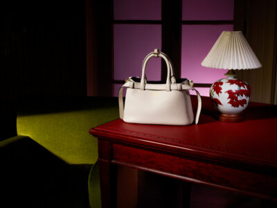 A stylish cream handbag rests on a polished red wooden table, paired with a decorative lamp featuring red leaf patterns. The scene is set against a purple-lit backdrop, creating a sophisticated ambiance. To the left, a green upholstered sofa adds contrast to the elegant composition, with light casting gentle shadows across the room's elements.