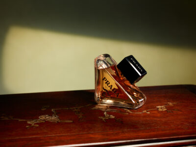 A perfume bottle rests on a polished wooden surface, exuding elegance and luxury. The bottle is angular, featuring a gold-toned liquid inside that catches the light, giving it a warm glow. Its black square cap contrasts with the rich, dark wood adorned with delicate, intricate carvings of birds and branches, set against a softly blurred background.