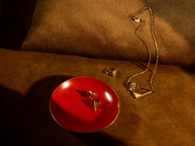 A luxurious composition features elegant gold jewelry with rose motifs, including a necklace and rings, elegantly placed on a rich brown fabric. A striking red plate holds a couple of the rings, enhancing their shine. The warm lighting and soft shadows create an inviting and sophisticated ambiance, emphasizing the intricate details and craftsmanship of each piece.
