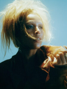 A person with light blonde, voluminous hair gazes thoughtfully upwards. Their face is softly illuminated, casting delicate shadows across their features. The background is a gradient of pale blue, creating an ethereal atmosphere. They're wearing a dark, textured outfit, with one hand partially visible, lightly poised against the chest. The composition conveys an introspective and serene mood.